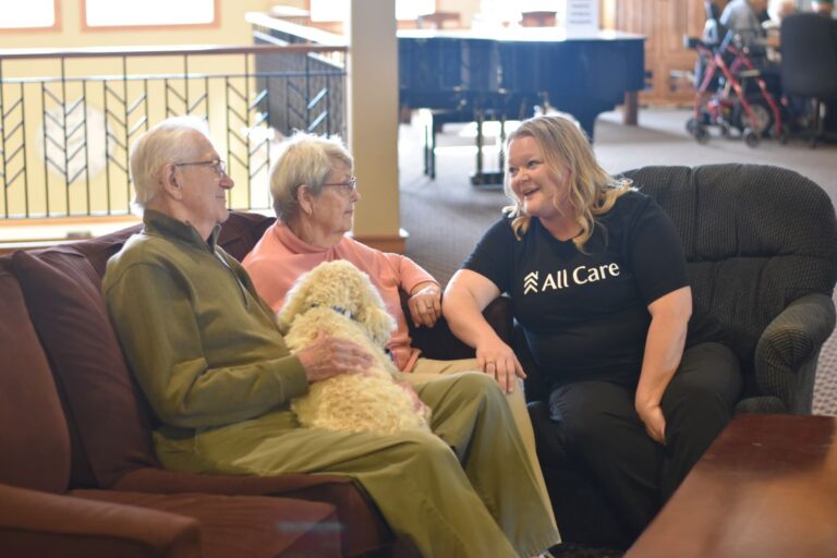 senior male and female seated on couch with white dog next to female All Care Staff.