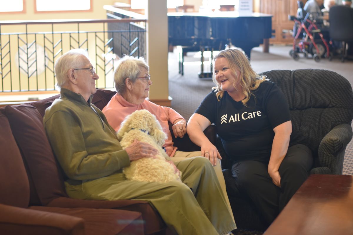 senior male and female seated on couch with white dog next to female All Care Staff.