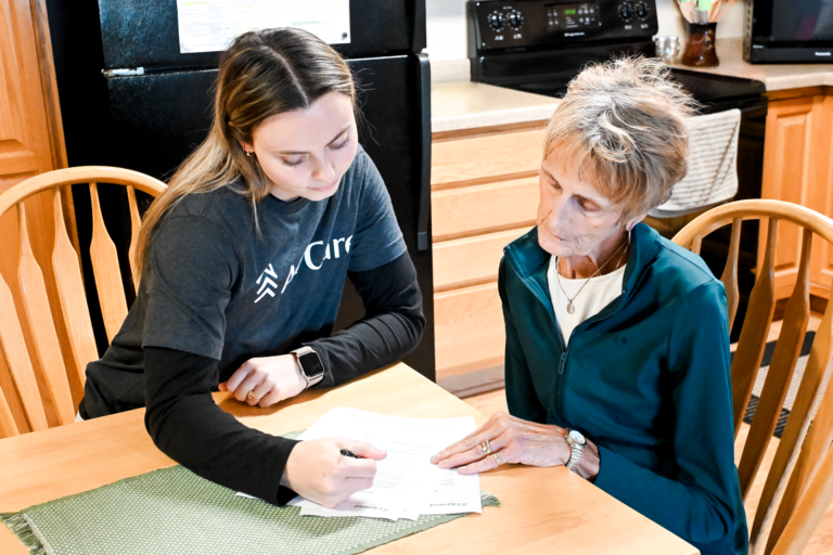 An All Care team member reviews paperwork with an older woman at her kitchen table.