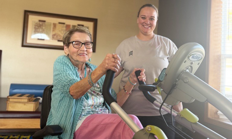 A senior woman holding the handlebars of a therapy machine while a female therapist stands closely behind her, assisting with the exercise.